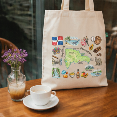 A cream-colored canvas tote bag with a colorful illustrated graphic of the Dominican Republic, displayed on a wooden table next to a white coffee cup and a vase of purple flowers.